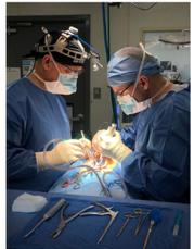 Two surgeons in scrubs and surgical loupes operating, with an instrument tray in the foreground